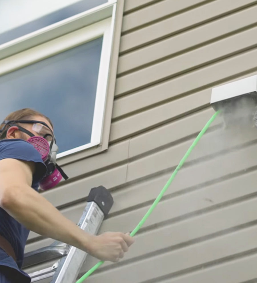 Technicien portant un masque de protection, nettoyant une sortie de ventilation extérieure sur le revêtement d'une maison, illustrant l'entretien professionnel des conduits de sécheuse ou de ventilation.